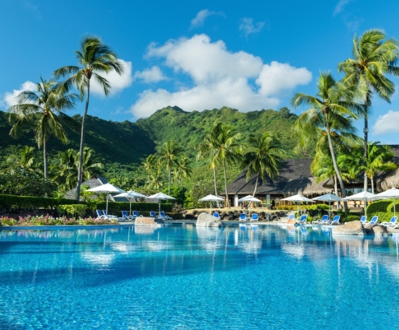 A bright blue resort swimming pool with lounge chairs and umbrellas, surrounded by lush green palm trees and a tropical mountain backdrop under a clear blue sky.