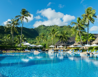 A bright blue resort swimming pool with lounge chairs and umbrellas, surrounded by lush green palm trees and a tropical mountain backdrop under a clear blue sky.