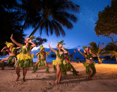 A group of dancers in traditional grass skirts and floral headdresses perform a Polynesian dance on a beach at dusk under a star-filled sky.