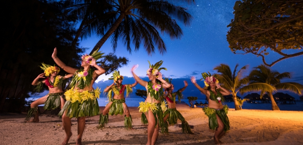A group of dancers in traditional grass skirts and floral headdresses perform a Polynesian dance on a beach at dusk under a star-filled sky.