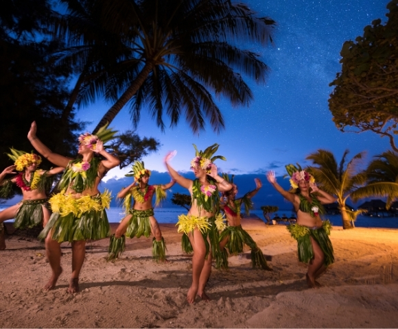 A group of dancers in traditional grass skirts and floral headdresses perform a Polynesian dance on a beach at dusk under a star-filled sky.