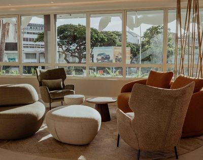 Sunlit modern lounge with sculptural beige and rust armchairs, ottomans, and a low table facing broad windows with leafy city views; calm, inviting mood.