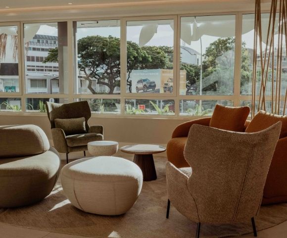 Sunlit modern lounge with sculptural beige and rust armchairs, ottomans, and a low table facing broad windows with leafy city views; calm, inviting mood.