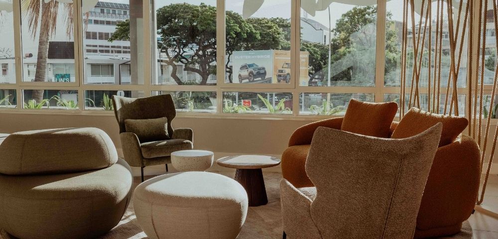 Sunlit modern lounge with sculptural beige and rust armchairs, ottomans, and a low table facing broad windows with leafy city views; calm, inviting mood.