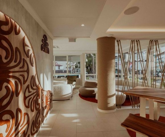 A serene modern lounge features a bold carved wooden wall, woven column, rope-like hanging decor, curved seating, and bright windows overlooking the city.