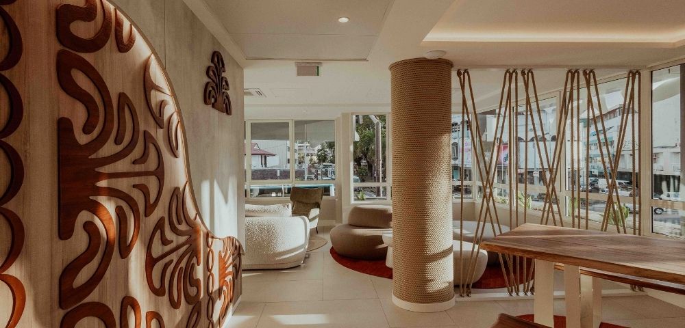 A serene modern lounge features a bold carved wooden wall, woven column, rope-like hanging decor, curved seating, and bright windows overlooking the city.