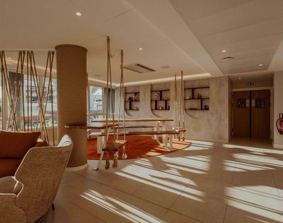 Sunlit hotel lounge with woven armchairs, sculptural wooden tables, curved shelving, and warm earth tones; quiet, inviting atmosphere.