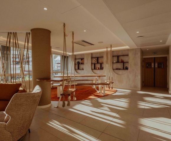Sunlit hotel lounge with woven armchairs, sculptural wooden tables, curved shelving, and warm earth tones; quiet, inviting atmosphere.