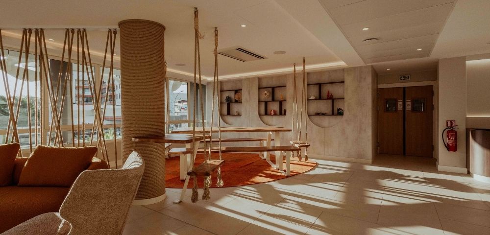 Sunlit hotel lounge with woven armchairs, sculptural wooden tables, curved shelving, and warm earth tones; quiet, inviting atmosphere.