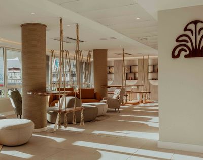 Sunlit modern lounge with sculptural woven columns, hanging rope chairs, curved sofas, and built-in shelves; warm, calm decor evokes a relaxed resort atmosphere.