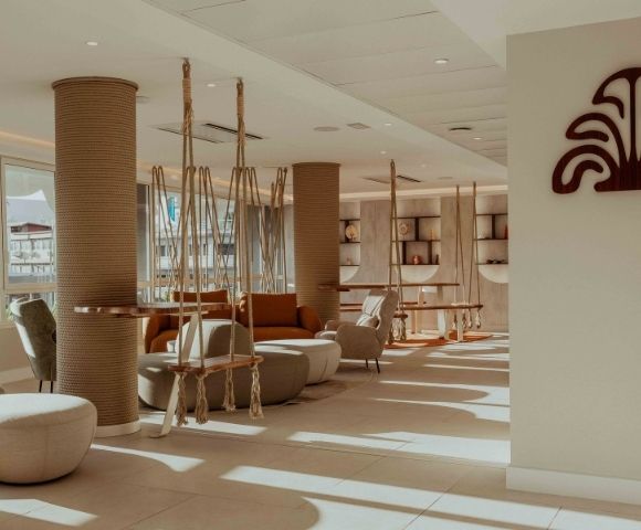 Sunlit modern lounge with sculptural woven columns, hanging rope chairs, curved sofas, and built-in shelves; warm, calm decor evokes a relaxed resort atmosphere.