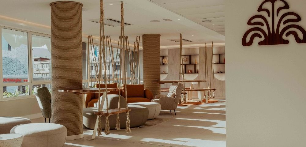 Sunlit modern lounge with sculptural woven columns, hanging rope chairs, curved sofas, and built-in shelves; warm, calm decor evokes a relaxed resort atmosphere.