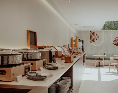 Sunlight fills a modern buffet with wooden counters, stainless serving pots, plated dishes, and empty tables beneath bold red wall motifs.