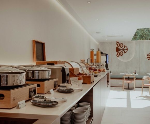 Sunlight fills a modern buffet with wooden counters, stainless serving pots, plated dishes, and empty tables beneath bold red wall motifs.
