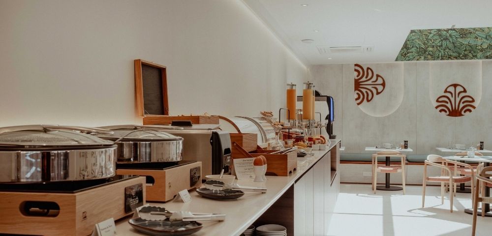 Sunlight fills a modern buffet with wooden counters, stainless serving pots, plated dishes, and empty tables beneath bold red wall motifs.