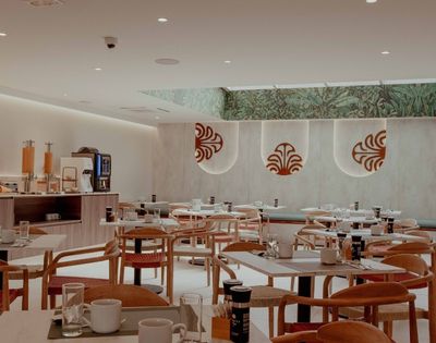Bright, modern restaurant dining room with neatly set tables, wooden chairs, a buffet counter, and red motifs on pale walls; calm, welcoming atmosphere.