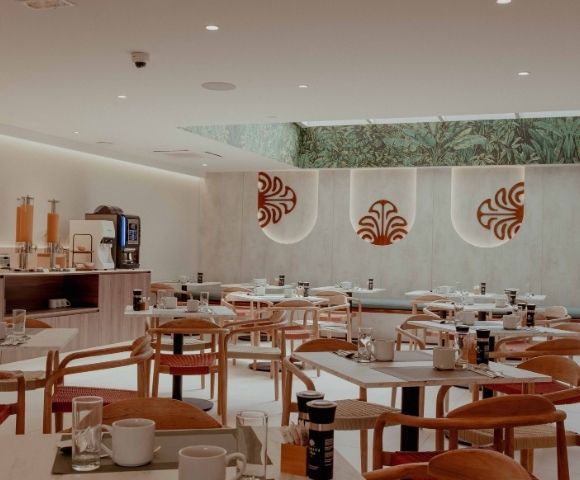 Bright, modern restaurant dining room with neatly set tables, wooden chairs, a buffet counter, and red motifs on pale walls; calm, welcoming atmosphere.