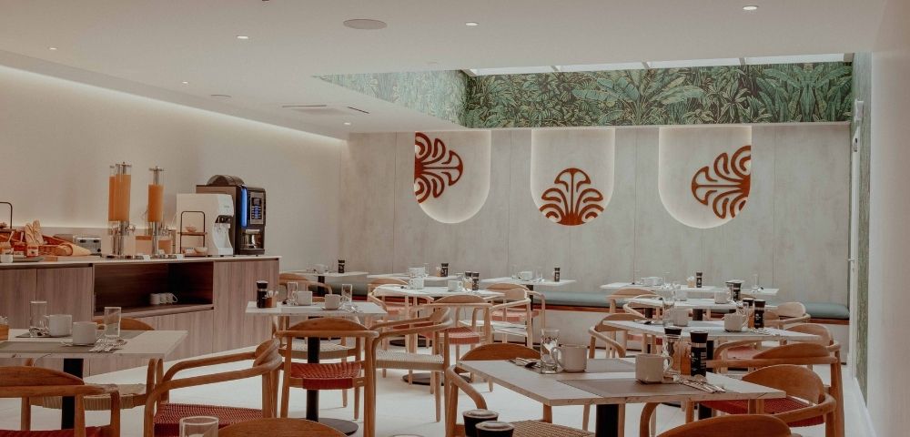 Bright, modern restaurant dining room with neatly set tables, wooden chairs, a buffet counter, and red motifs on pale walls; calm, welcoming atmosphere.