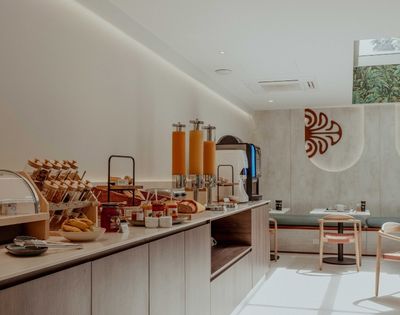 A modern breakfast buffet with pastries, bread, orange juice dispensers, and dining tables; warm tones and decorative wall art create a calm, inviting mood.