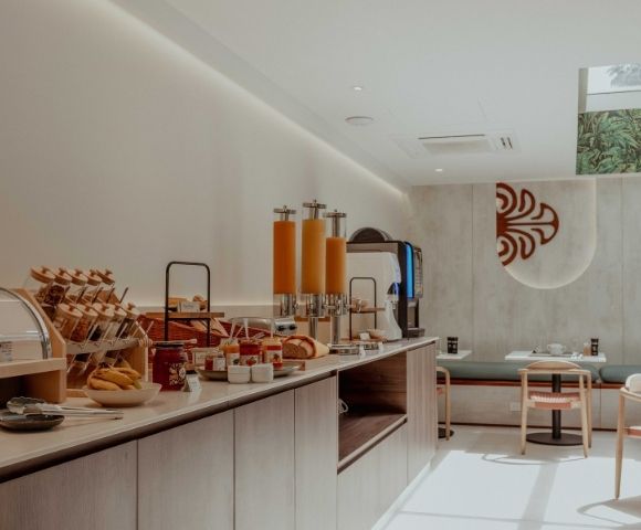 A modern breakfast buffet with pastries, bread, orange juice dispensers, and dining tables; warm tones and decorative wall art create a calm, inviting mood.