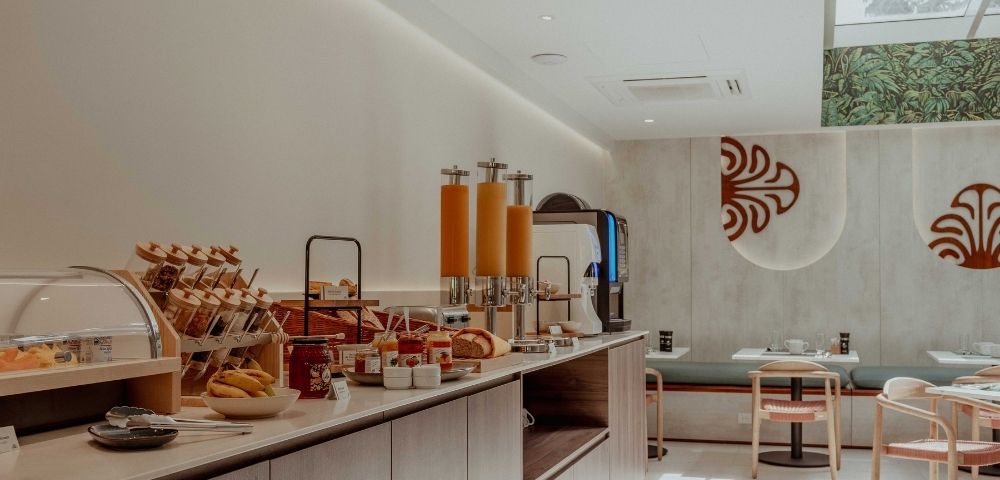 A modern breakfast buffet with pastries, bread, orange juice dispensers, and dining tables; warm tones and decorative wall art create a calm, inviting mood.