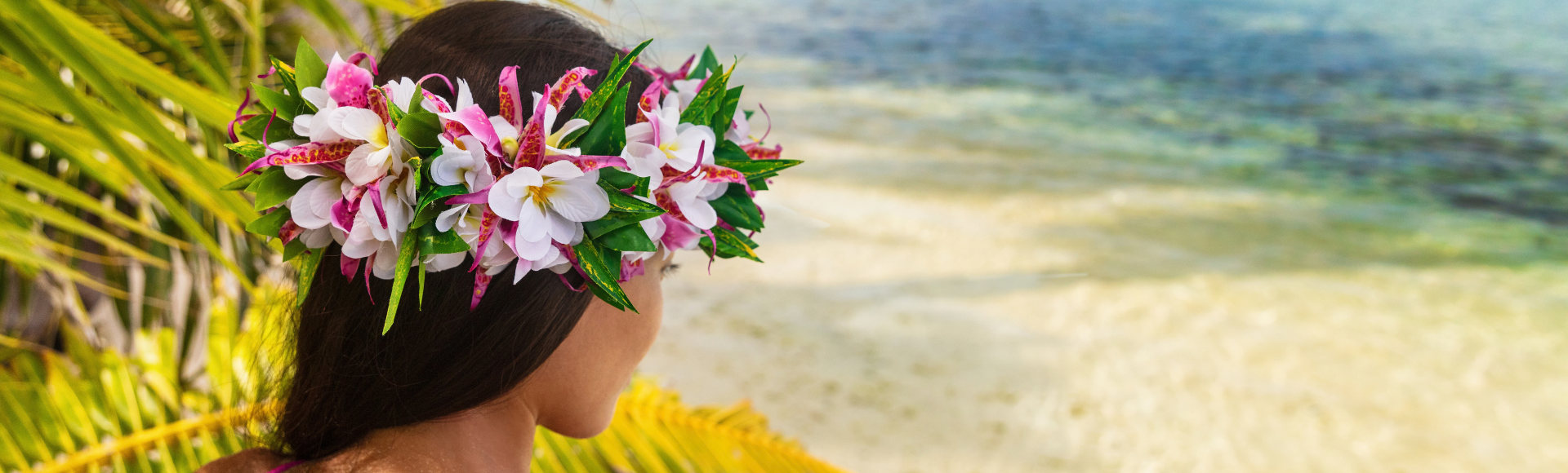 A person wearing a colorful floral crown looks out toward a clear turquoise lagoon, framed by arching palm fronds in the foreground. The tropical shoreline, bright greenery, and calm ocean water create a peaceful island setting under soft daylight.