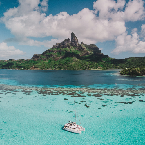 Catamaran anchored in turquoise lagoon waters beneath a lush, jagged volcanic island and partly cloudy blue sky.