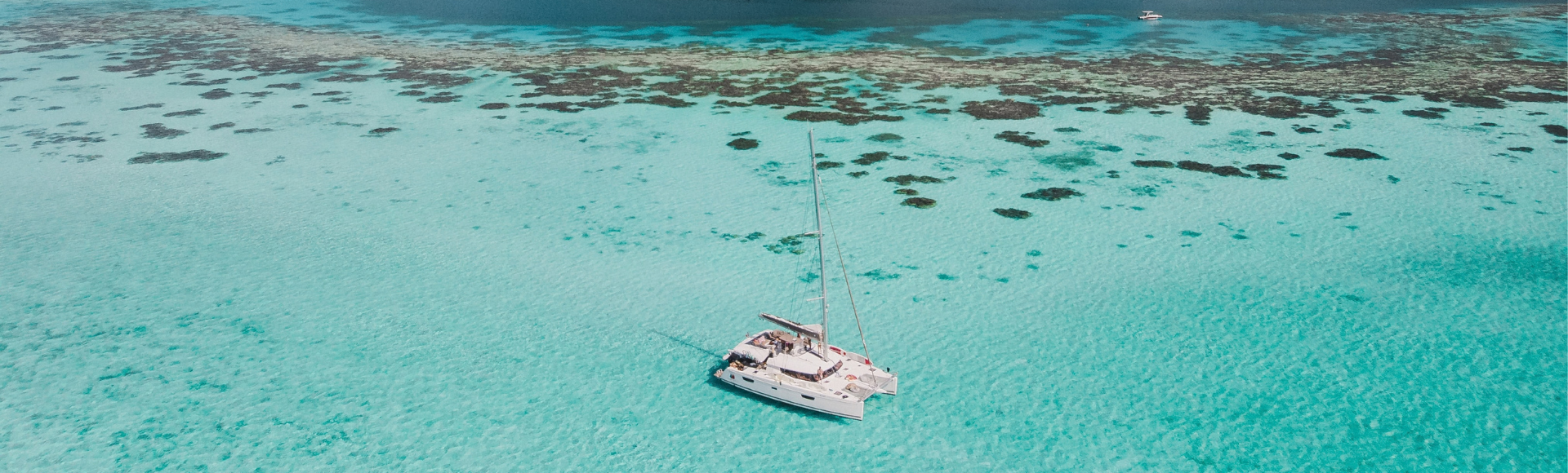 Catamaran anchored in turquoise lagoon waters beneath a lush, jagged volcanic island and partly cloudy blue sky.