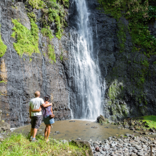 Two hikers stand beside a rocky pool beneath a tall waterfall cascading down a moss-covered cliff.