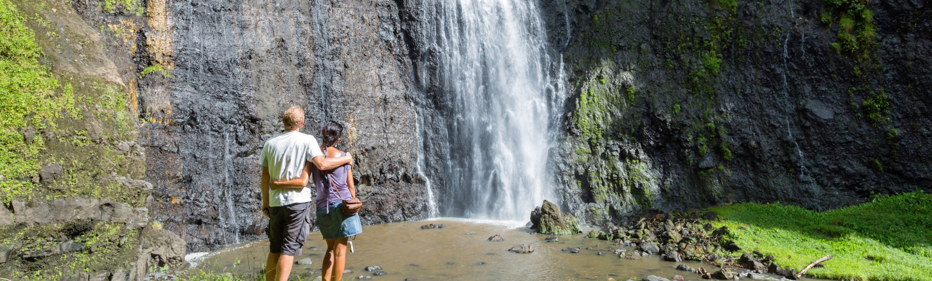 Two hikers stand beside a rocky pool beneath a tall waterfall cascading down a moss-covered cliff.