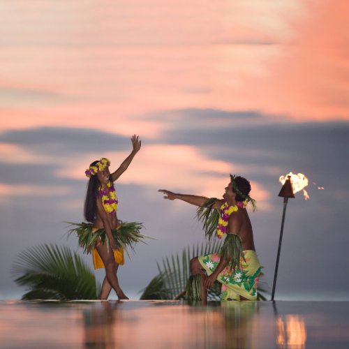 Two Polynesian dancers in floral leis and grass skirts perform at sunset beside a flaming torch, reflected in shallow water.