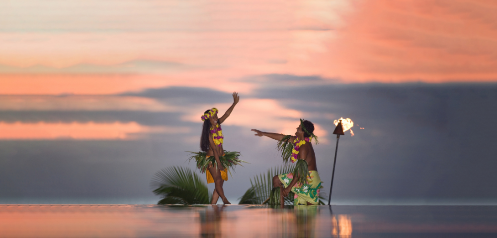Two Polynesian dancers in floral leis and grass skirts perform at sunset beside a flaming torch, reflected in shallow water.