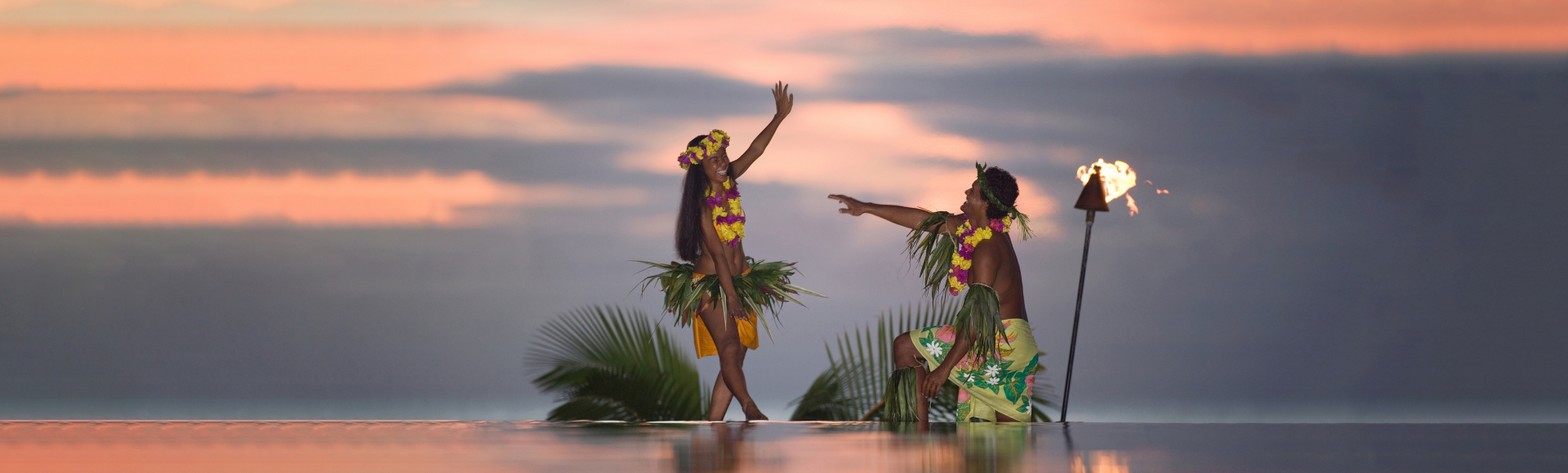 Two Polynesian dancers in floral leis and grass skirts perform at sunset beside a flaming torch, reflected in shallow water.
