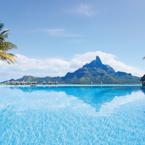 A bright blue infinity pool reflects a large, jagged mountain peak under a clear sky, with small thatched-roof bungalows visible in the distance.