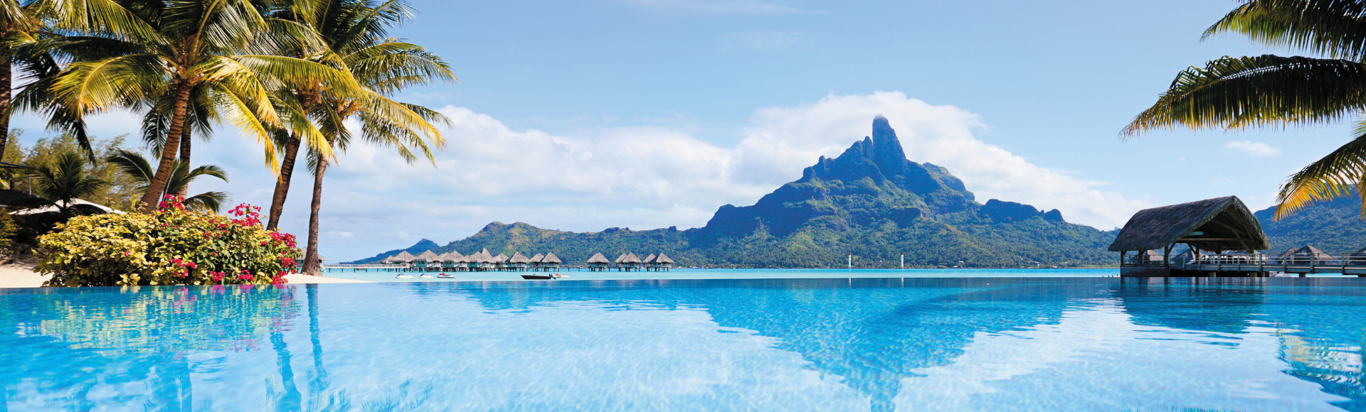 A bright blue infinity pool reflects a large, jagged mountain peak under a clear sky, with small thatched-roof bungalows visible in the distance.
