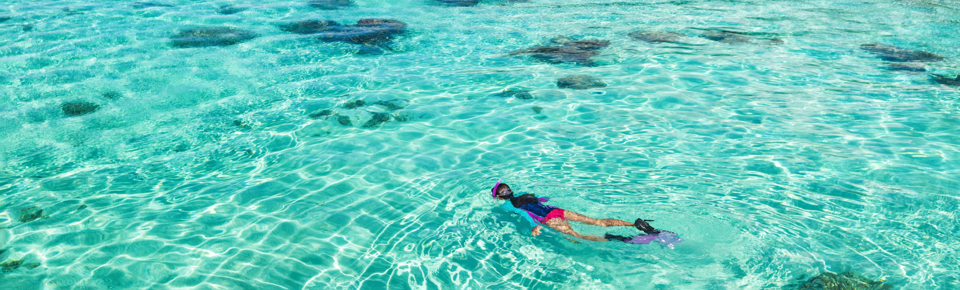 A person snorkeling in crystal-clear turquoise water with visible coral reefs below, set against a backdrop of lush green mountains under a blue sky.
