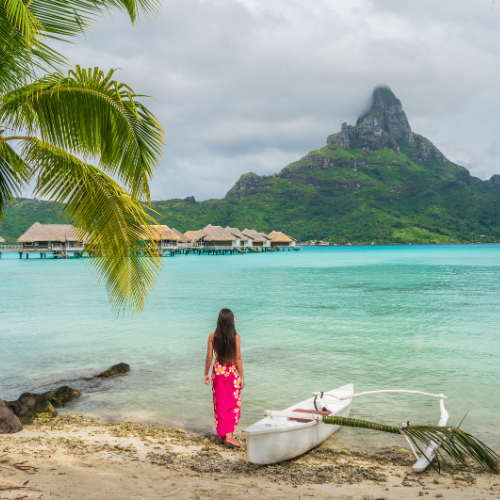 A tropical beach scene with clear turquoise water, a white outrigger canoe resting on the sandy shore, and a person standing at the water’s edge facing a lush green mountain island. Palm fronds frame the foreground, while overwater bungalows stretch across the lagoon beneath a cloudy sky.