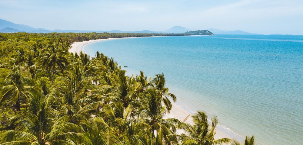A sweeping aerial view of a tropical coastline featuring a crescent-shaped white sand beach lined with lush green palm trees. The turquoise ocean stretches to the horizon under a clear blue sky, with distant hazy mountains.