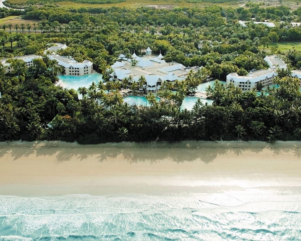 Aerial view of a luxurious resort with white buildings and multiple swimming pools nestled amidst lush green palm trees and dense tropical foliage, bordering a wide sandy beach and the turquoise ocean.