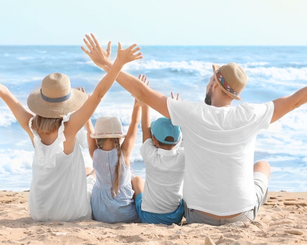 A family of four, including two adults and two children, sits on a sandy beach with their arms raised in celebration, facing the ocean and a bright blue sky.