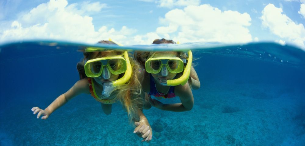 Two young children wearing yellow snorkeling masks and snorkels swim underwater in a clear blue ocean, with the surface and clouds visible above.
