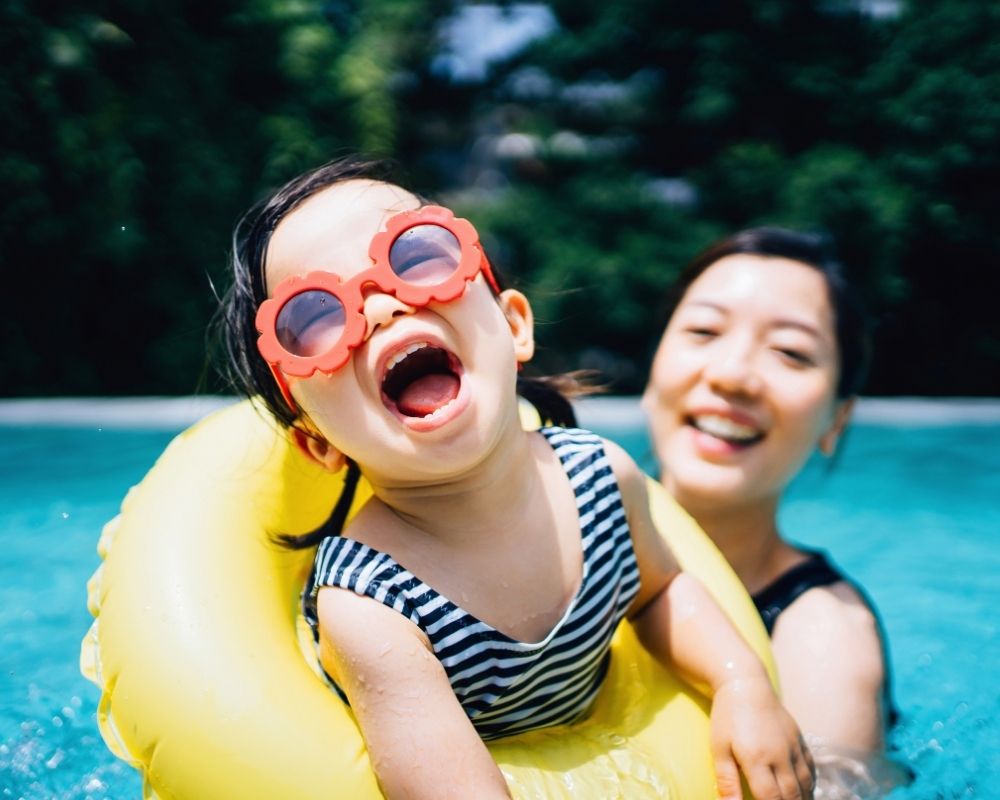 A child wearing large red flower-shaped sunglasses and a striped swimsuit is joyfully laughing while floating in a bright yellow swim ring in a pool, with an adult smiling behind them.
