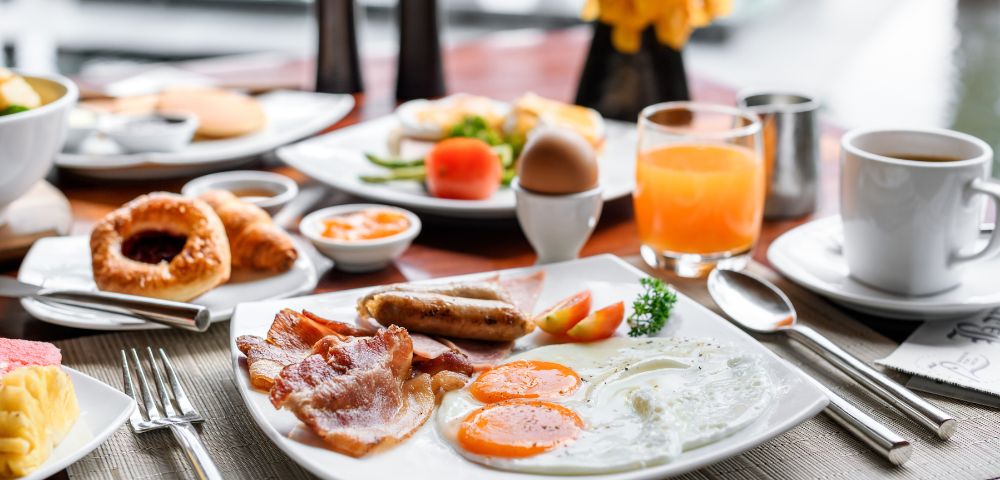 A hearty breakfast plate featuring fried eggs, crispy bacon, and sausages, served with a glass of orange juice, a boiled egg in a holder, and pastries on the side.