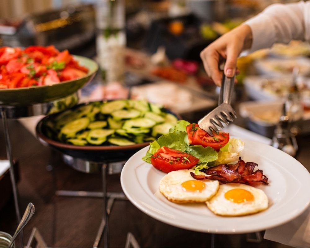 A person is serving a plate of breakfast with two fried eggs, bacon, and a salad topped with tomatoes and lettuce from a buffet. In the background are bowls of sliced tomatoes and cucumbers.