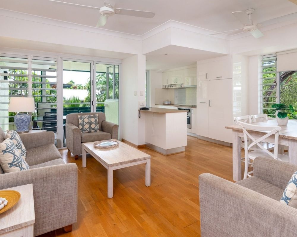 A modern, open-plan living and dining area with a kitchen in the background, featuring light-colored furniture, wooden floors, and large windows with views of lush greenery.