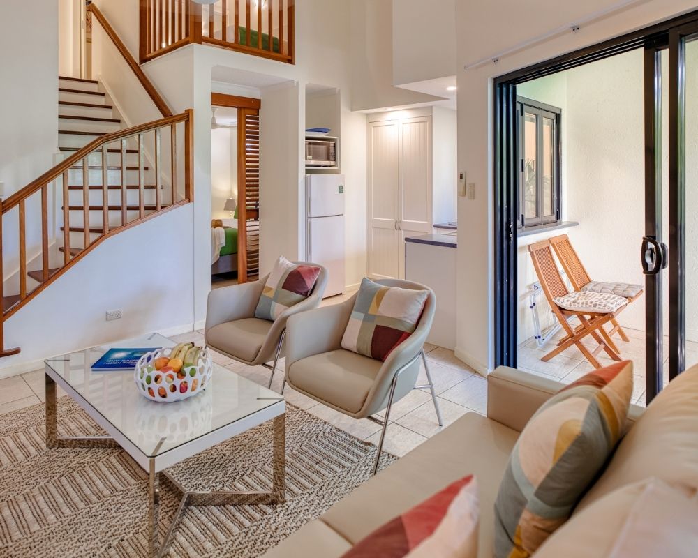 A bright, modern living area with a multi-level design. A wooden staircase ascends to the left, leading to an upper level. In the center, two light-colored armchairs with patterned throw pillows face a glass-topped coffee table holding a book and a fruit basket. A sofa is partially visible in the foreground. To the right, a sliding glass door opens to a small patio with two wooden chairs. A small kitchen area with a refrigerator and cabinets is visible in the background.