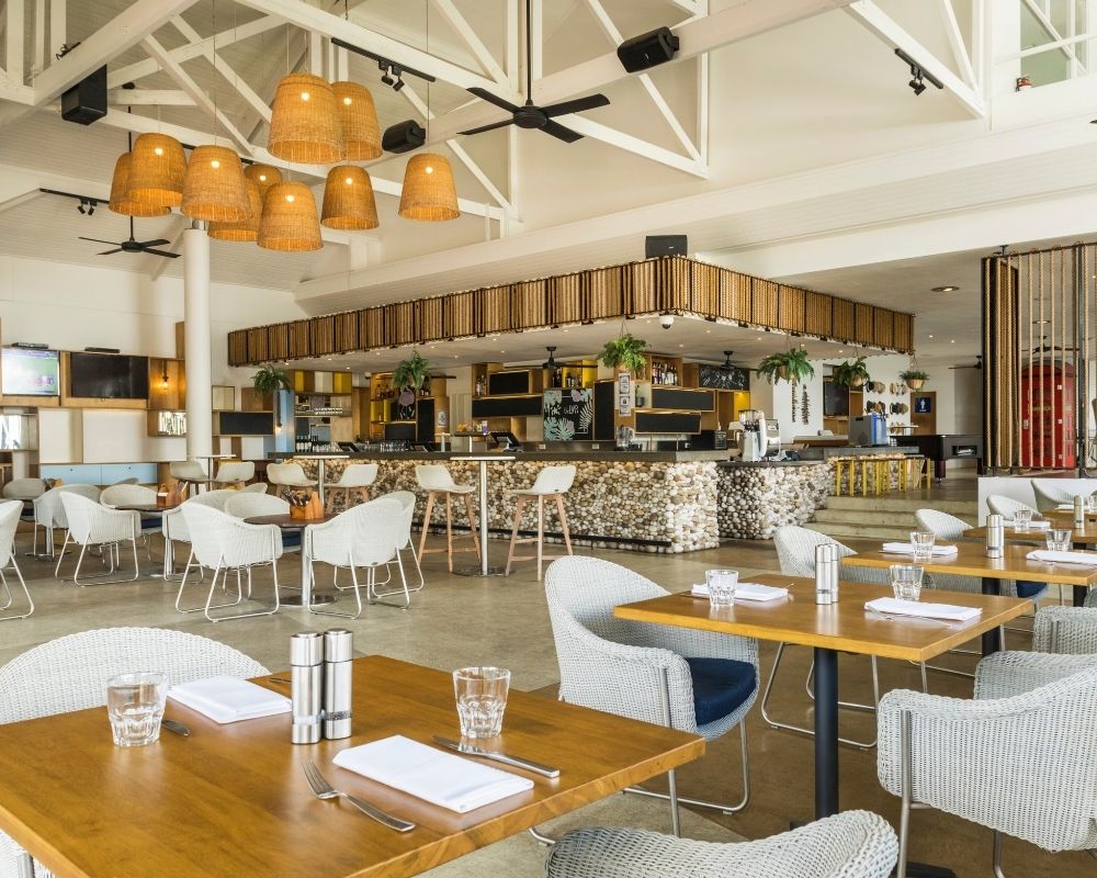 A bright, modern restaurant with a large bar and dining area, featuring natural wood accents, woven light fixtures, and white wicker chairs.
