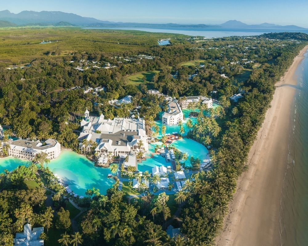 Aerial view of a luxurious resort complex with expansive turquoise pools surrounded by lush greenery and palm trees, bordering a sandy beach and the ocean.