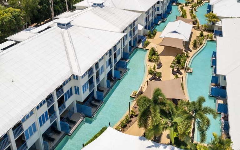 Aerial view of a resort with long white buildings featuring blue balconies and swimming pools winding between them, surrounded by lush green trees.