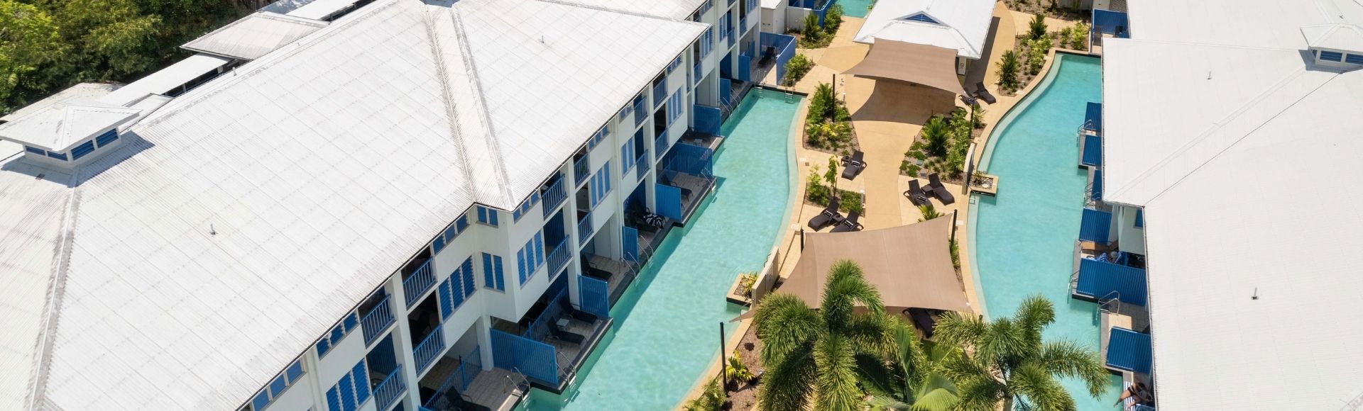 Aerial view of a resort with long white buildings featuring blue balconies and swimming pools winding between them, surrounded by lush green trees.